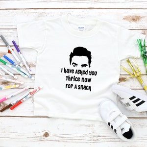 May include: A white short-sleeved t-shirt featuring a black graphic of a person's eyes and hair above the text "I have Asked you Thrice now FOR A SNACK" in a bold, black, rounded font. The shirt is displayed with colorful markers and shoes.