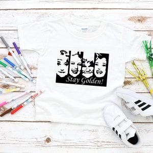 May include: White t-shirt with a black and white graphic of four faces and the text "Stay Golden!"