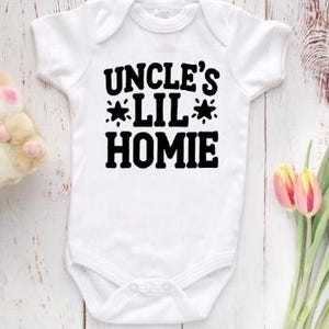 Peut inclure: Combinaison blanche pour bébé avec un texte noir qui dit "Uncle's Lil' Homie" avec une étoile de chaque côté de "Lil".