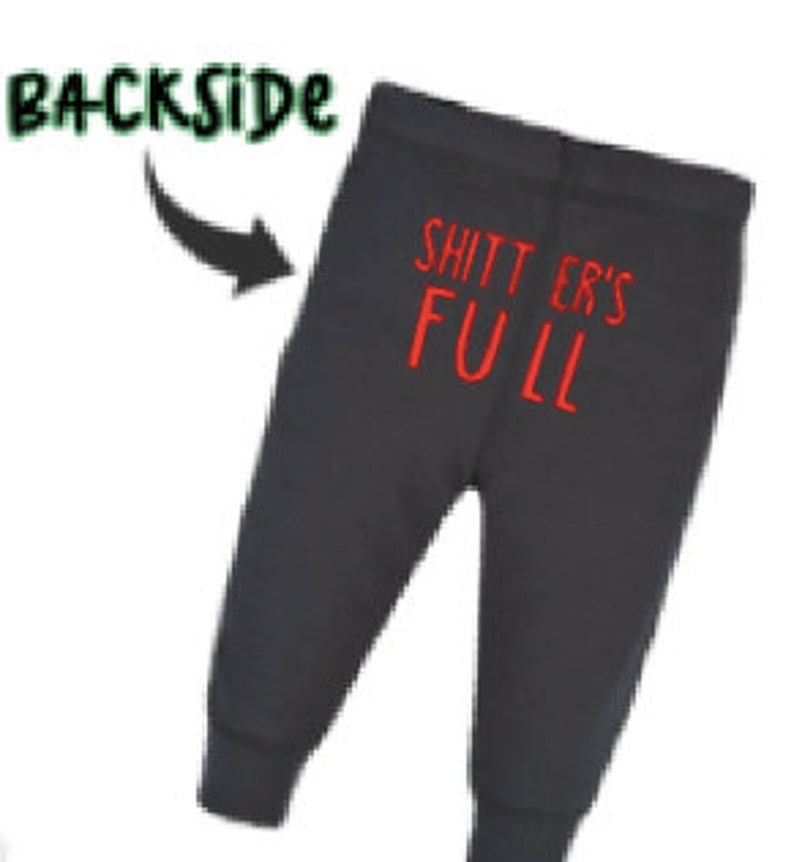 May include: Black sweatpants with the text "SHITTER'S FULL" in red on the back. The word "BACKSIDE" is written in green above the sweatpants with an arrow pointing to the text. The sweatpants have a fitted ankle.