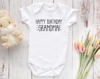 Könnte beinhalten: Weißer Baby Strampler mit schwarzem Text, der "Happy Birthday Grandma!" lautet. Der Strampler liegt auf einem weißen Holz Hintergrund mit einem Stofftier Hasen und rosa Tulpen.