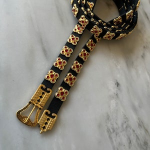 May include: A black leather belt with a gold buckle and red jeweled accents. The belt is adorned with a repeating pattern of gold squares with red jewels in the center.