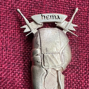 May include: A silver-colored metal pin featuring a helmet design with crossed swords and a banner that reads "hema". The pin has a detailed, textured surface and is likely a collectible or decorative item.