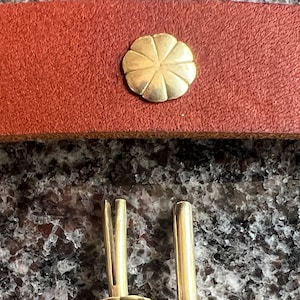 Puede incluir: Dos remaches de metal dorado con una cabeza decorativa en forma de flor. Los remaches están sobre una superficie marrón texturizada.