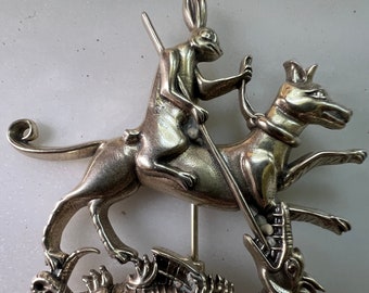 Puede incluir: Un broche plateado que representa un conejo montando un perro, que está atacando a un dragón. El conejo sostiene una lanza en su mano derecha.