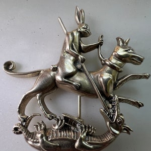 May include: A silver-toned brooch depicting a rabbit riding a dog, which is attacking a dragon. The rabbit holds a spear in its right hand.