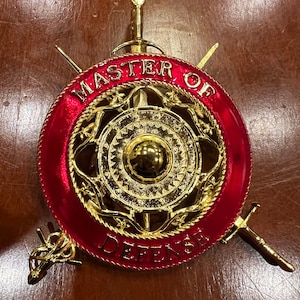 Pode incluir: Um escudo de metal dourado com fundo vermelho e as palavras "MASTER OF DEFENSE" ao redor da borda. O escudo tem um design em relevo de uma espada e um círculo com um círculo menor no centro.