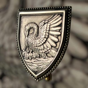 May include: A silver-toned shield-shaped brooch with a detailed relief of a bird with outstretched wings, possibly a pelican, holding small figures. The brooch has a beaded border and a gold-colored pin.