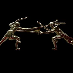 Pode incluir: Broche de metal dourado que retrata duas figuras envolvidas numa luta de espadas. As figuras estão em poses dinâmicas, com espadas levantadas e estendidas. O fundo é preto sólido.