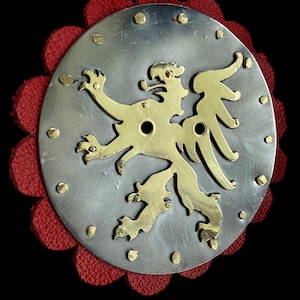 May include: A circular brooch featuring a silver-toned metal base with a gold-coloured lion emblem. The brooch is embellished with small gold rivets and set on a red, scalloped leather backing. The design is set against a black background.
