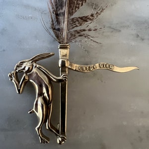 May include: A gold-colored metal bookmark featuring a hare holding a banner with the words "carpe diem." A feather is attached to the top of the bookmark. The background is a light gray and white marble surface.