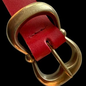 Puede incluir: Un cinturón de cuero rojo con una hebilla dorada. La hebilla tiene un diseño curvo único. El cinturón está en ángulo sobre el marco contra un fondo negro, destacando el contraste entre el cuero rojo y la hebilla dorada.