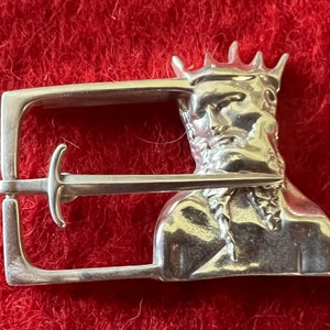Puede incluir: Hebilla de cinturón de plata con un diseño detallado de la cabeza de un hombre coronado con una espada que pasa a través de la hebilla.