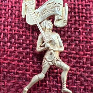 May include: A gold-colored metal figurine of a person running with a sword and a banner that reads "Veni Vidi Vici".
