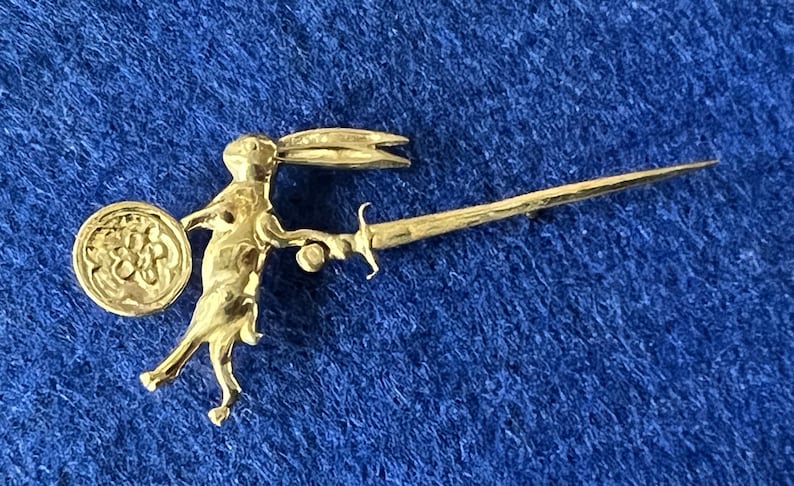May include: A gold-colored brooch featuring a rabbit holding a sword and a shield with a floral design.