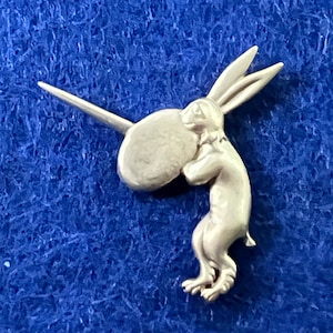 Puede incluir: Una figurita de conejo de color plateado que sostiene un objeto redondo. El conejo tiene orejas largas y está de pie sobre sus patas traseras.