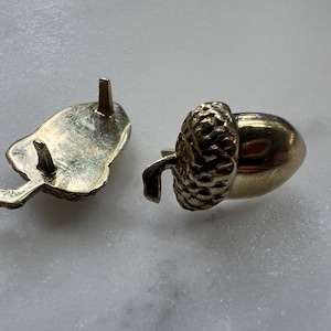 May include: Two gold-colored acorn-shaped objects. One is a complete acorn, the other is the acorn cap. The acorn has a textured cap and a smooth, oval body. The cap has three small posts for attachment. The objects are likely decorative or jewelry components.