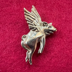 May include: Silver pig charm with wings, a whimsical and detailed design. The pig is flying with its wings spread out, and its snout is pointed forward. The charm is perfect for adding a touch of whimsy to any jewelry piece.