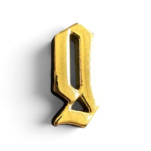 May include: A gold-colored metal pendant in the shape of the letter Q, rendered in a stylized gothic typeface. The piece features a polished, reflective surface with sharp, angular edges and a hollow center, creating a bold, three-dimensional appearance.