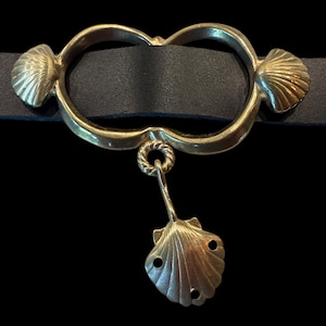 May include: A black leather belt with a gold-colored metal buckle. The buckle features a double-loop design with seashell accents and a dangling seashell pendant. The metal has a vintage, antique appearance.