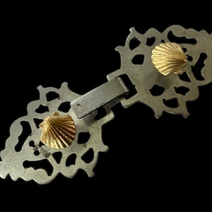 May include: Ornate silver-toned metal clasp with a decorative openwork design. The clasp features two gold-colored seashell-shaped accents. The clasp is designed to fasten two pieces of fabric or material together.