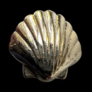 Op de afbeelding: Een gedetailleerde, metalen zeeschelp met een getextureerd oppervlak. De schelp heeft een lichtgouden kleur en heeft een geribbeld patroon. De achtergrond is effen zwart, wat de vorm en details van de schelp benadrukt. Dit decoratieve stuk kan worden gebruikt als een sieradencomponent of woondecoratie.