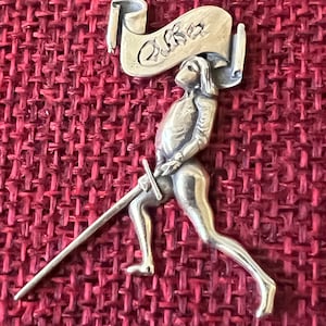 Puede incluir: Una figurita de plata de un caballero sosteniendo una espada y un rollo con la palabra "Cura" escrita en él. La figurita está sobre un fondo rojo texturizado.