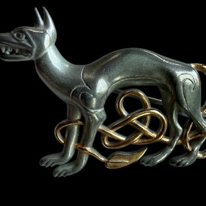 May include: A silver metal dog figurine with gold-coloured Celtic knot accents. The dog is in a stylized pose with its mouth open, and the knotwork forms a decorative element around its legs.