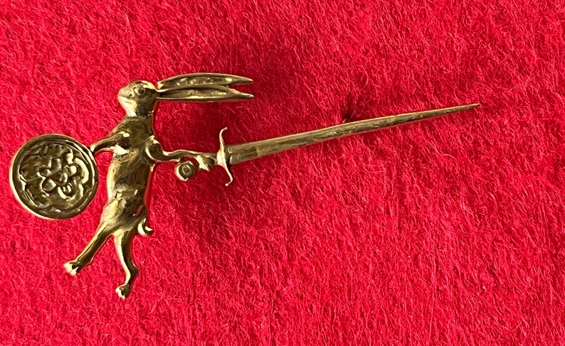 May include: A gold-colored rabbit figurine holding a sword and shield. The rabbit is standing on a red surface.