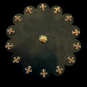 Könnte beinhalten: Eine kreisförmige schwarze Ledermatte mit gewelltem Rand, die mit zwölf metallisch goldfarbenen Fleur-de-Lis-Emblemen am Rand und einem einzelnen metallisch goldfarbenen blumenförmigen Niet in der Mitte der Matte verziert ist.