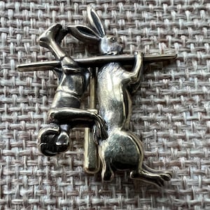 May include: A gold-toned metal pin depicting a rabbit being speared by a man. The rabbit is standing on its hind legs and the man is hanging upside down.