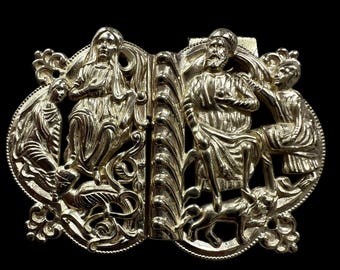 Puede incluir: Una hebilla de metal dorado con diseños intrincados que representan figuras y animales. La hebilla está hecha de dos piezas que encajan entre sí.
