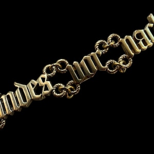 May include: A gold-colored necklace with the words "Landes und Macht" in gothic lettering. The chain links are intricately designed, and the background is black.