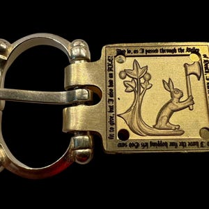 May include: A metal belt buckle with a silver and gold finish. The buckle features a rectangular plate with an engraved image of a rabbit holding an axe, with text. The buckle is designed with a D-ring and a hinged bar.