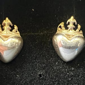 May include: A pair of silver-toned heart-shaped earrings with gold-colored crown details. The earrings are displayed against a dark background, showcasing their reflective surfaces and intricate design. These earrings are a stylish accessory.