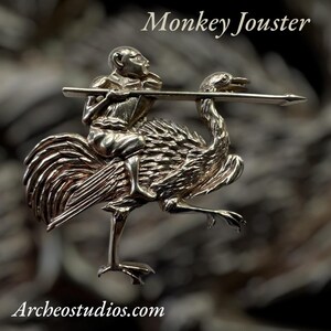May include: A silver-toned brooch depicting a monkey riding an ostrich, holding a lance. The brooch is detailed with intricate textures and features the text "Monkey Jouster" and "Archeostudios.com".