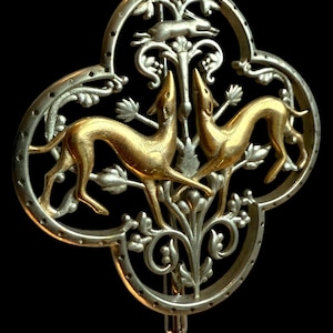 May include: Ornate silver and gold-toned brooch featuring two greyhounds facing each other, with a running hare above. The brooch has a quatrefoil shape with intricate floral and foliate details. The background is black.