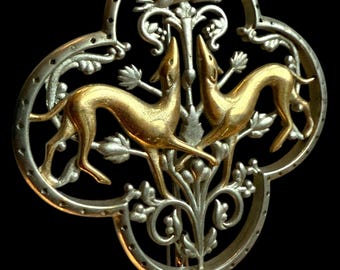 Puede incluir: Broche ornamentado en tonos plateados y dorados, con dos galgos enfrentados y una liebre corriendo encima. El broche tiene forma de cuadrifolio con intrincados detalles florales y foliares. El fondo es negro.