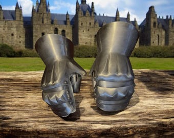 May include: A pair of silver metal gauntlets with a textured surface and pointed fingers. The gauntlets are sitting on a wooden surface.