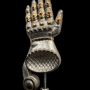Puede incluir: Escultura de mano de metal plateado con detalles dorados en los nudillos y la muñeca. La mano está en posición abierta, montada sobre un soporte plateado sobre un fondo negro. La escultura tiene un diseño detallado y texturizado.