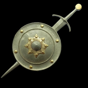 以下が含まれることがあります： 金色の星型の中心と金のスタッドが付いた、丸い盾のデザインの銀色のブローチ。 剣が盾を斜めに横切って配置されています。 ブローチの裏側には、長く尖ったピンが付いています。