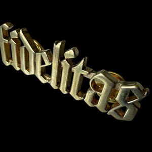 May include: A gold-toned metal lapel pin featuring the word fidelitas in a raised, three-dimensional gothic-style font. The letters have a polished, reflective surface with sharp, angular edges, set against a plain black background.