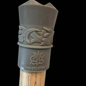 May include: A gray bottle stopper with a decorative floral design and the text "archeostudios.com". The stopper is on a wooden dowel. The stopper has a crown logo.