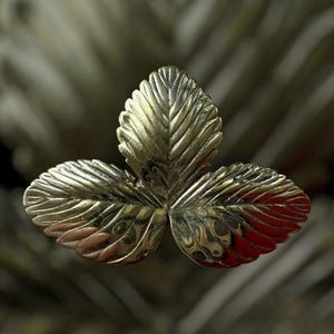 May include: A gold-coloured metal leaf brooch featuring three detailed leaves. The brooch has intricate veining and a textured surface, giving it a realistic look. The blurred background highlights the brooch's design and craftsmanship.