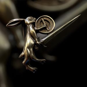 May include: A bronze-toned brooch featuring a standing hare holding a round shield and a long, pointed sword. The hare is detailed with visible fur and facial features. The brooch has a pin on the back for attachment.