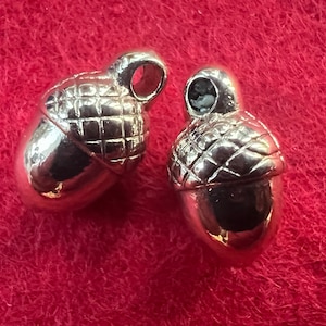 May include: Two silver acorn charms with a detailed textured design. The charms have a loop at the top for attaching to jewelry or other crafts.