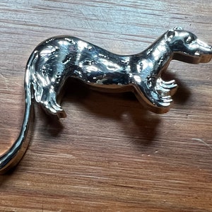 Puede incluir: Una figurita de metal de color plateado de una comadreja o un hurón. La figurita es detallada y tiene un aspecto realista.
