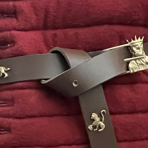 May include: A brown leather belt with a silver buckle featuring a crowned figure. The belt has three decorative metal accents: a wolf, a lion, and a crowned figure.