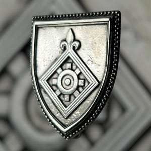 Puede incluir: Broche plateado en forma de escudo con un diseño detallado. El broche presenta una forma de diamante central con un patrón de sol, una flor de lis y un borde con cuentas. El metal tiene una apariencia ligeramente envejecida.
