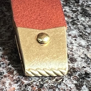 May include: Close-up of a brown leather strap with a gold metal accent and a gold rivet. The strap has a decorative braided edge.